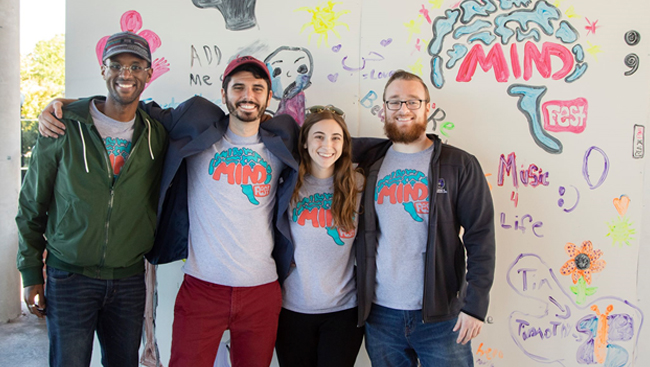 The primary members of the MIND Coalition, including cofounders Stallmann and Lovan, stand in front of an art wall at MINDfest 2018 that invited attendees to paint what mental health means to them. From left to right: Nathan Jones, Bradford Martins, Rebecca Stallmann, and Elijah Lovan.