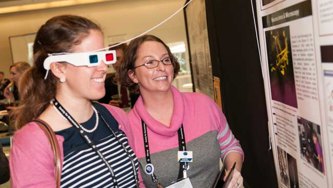 Two women engaged in a Brain Awareness activity. One of them wears 3D glasses while the other reads a poster.