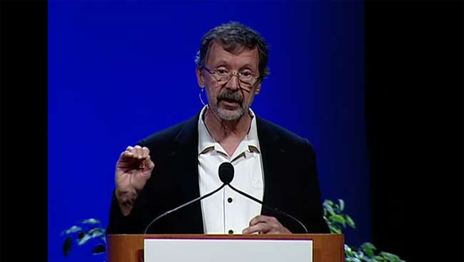 Image of Ed Catmull speaking at a microphone in front of a blue and black background. 