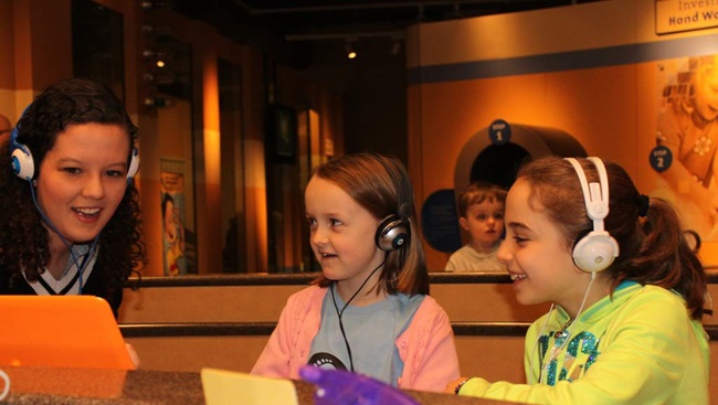 Two children are wearing headphones to participate in a science activity with a female neuroscientist.