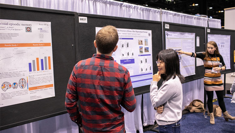 A women in a grey sweater presents her poster to a man in a red and grey plaid shirt at Neuroscience 2019