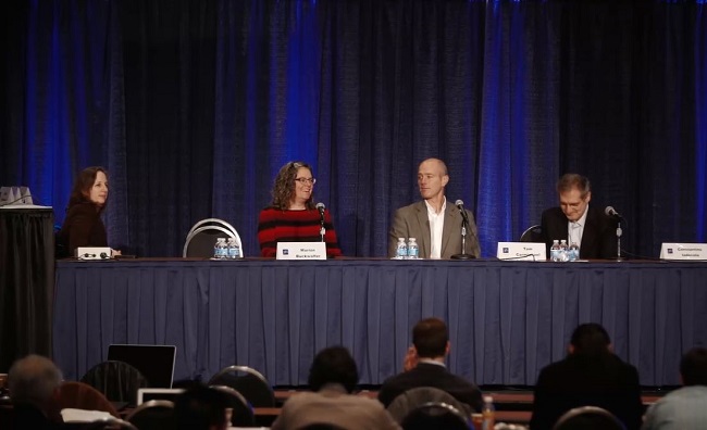 Panel of 5 speakers sitting a table in front of a blue curtain