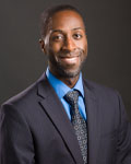 Headshot of Nii Addy wearing a blue suit in front of a gray backdrop.